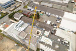 Drohnenfoto, Maschinenteil wird auf dem Produktionsgelände von Raps mit einem Kran durch das Dach eines Fabrikgebäudes gehoben