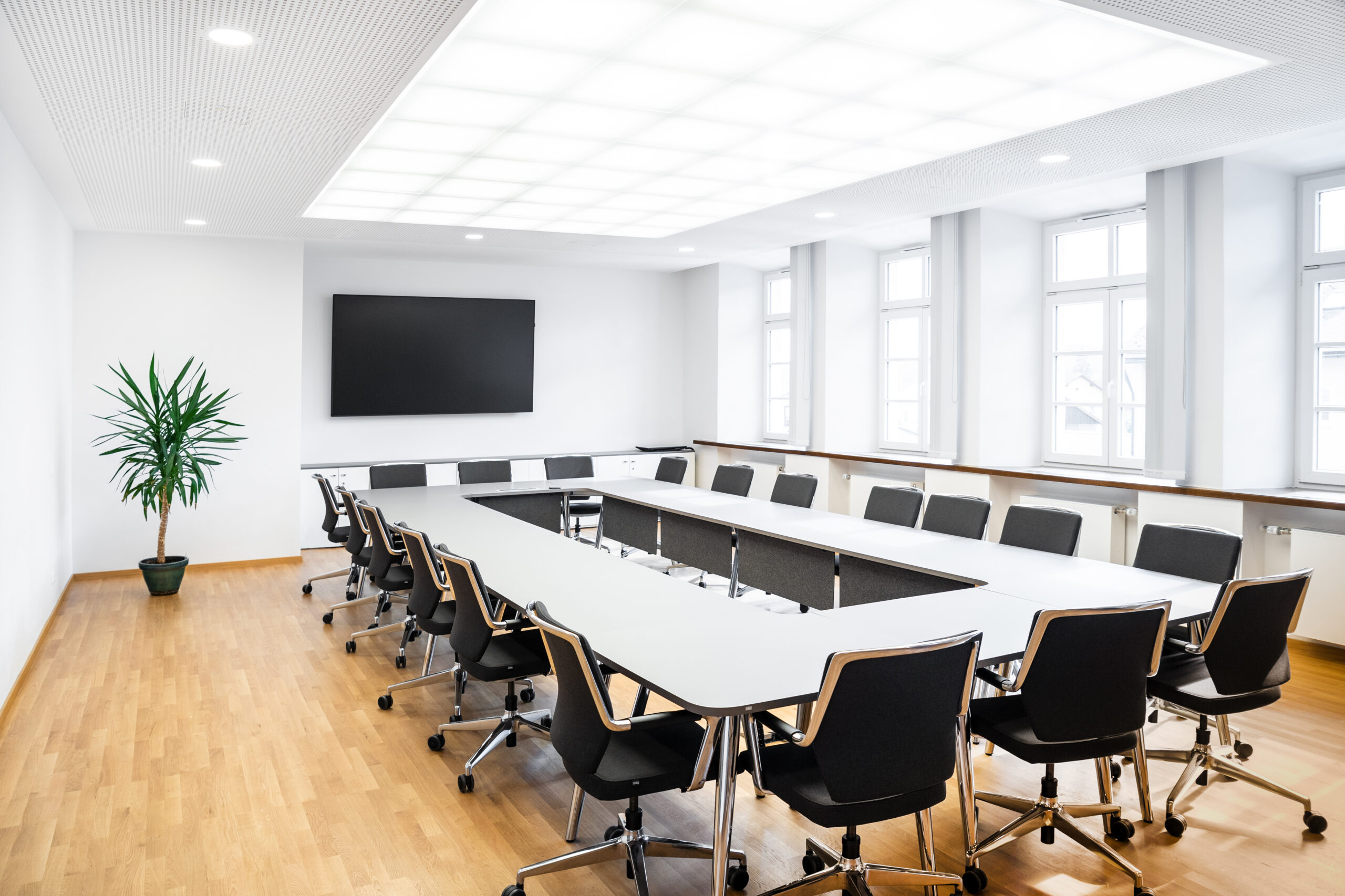 Moderner Besprechungsraum, Tische sind in der Raummitte o-förmig angeordnet, darum herum stehen Stühle, im Hintergrund hängt ein großer Bildschirm an der Wand