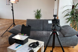 Interview Setup Fotostudio Kingunion, Links eine Dauerleuchte, in der Mitte ein Sofa, davor eine Kamera auf Stativ neben einem Tisch auf dem ein Filmklappe und Kopfhörer liegen