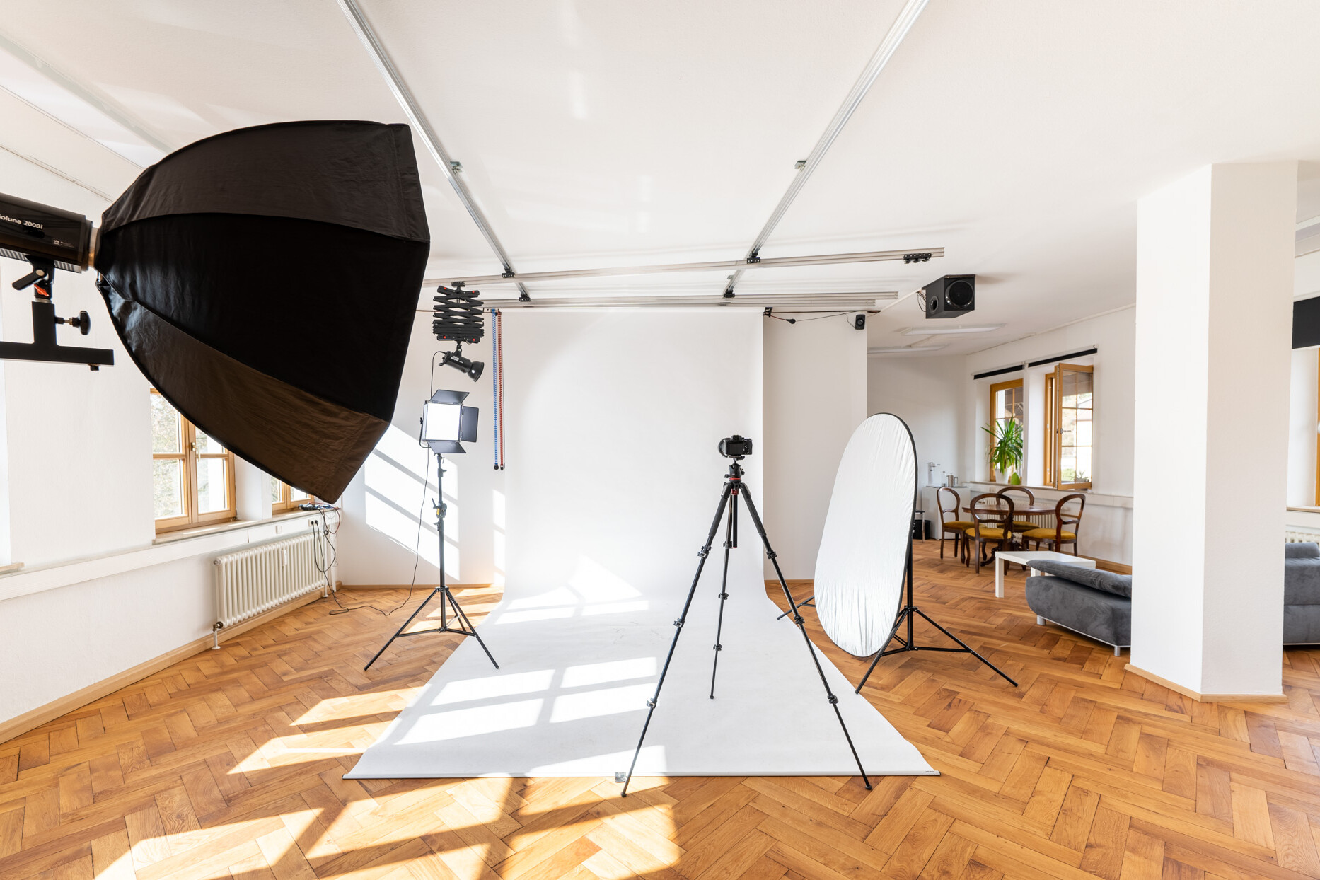 Fotostudio Kingunion, weißer Studiohintergrund ist ausgerollt, links ein LED-Dauerlicht mit Lightbox und eine Flächenleuchte, in der Raummitte eine Kamera auf Stativ, rechts ein Faltreflektor, darüber ein Deckenschienensystem
