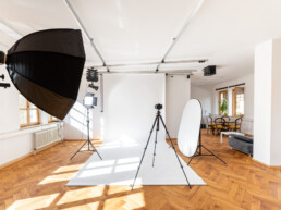 Fotostudio Kingunion, weißer Studiohintergrund ist ausgerollt, links ein LED-Dauerlicht mit Lightbox und eine Flächenleuchte, in der Raummitte eine Kamera auf Stativ, rechts ein Faltreflektor, darüber ein Deckenschienensystem
