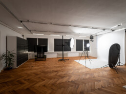 Fotostudio Kingunion, weißer Studiohintergrund ist ausgerollt, davor steht ein LED-Dauerlicht mit Lightbox und eine Flächenleuchte, eine Kamera auf Stativ, rechts ein Faltreflektor, darüber ein Deckenschienensystem