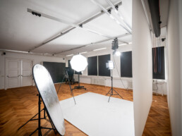 Fotostudio Kingunion, weißer Studiohintergrund ist ausgerollt, rechts ein LED-Dauerlicht mit Lightbox und eine Flächenleuchte, in der Raummitte eine Kamera auf Stativ, links ein Faltreflektor, darüber ein Deckenschienensystem